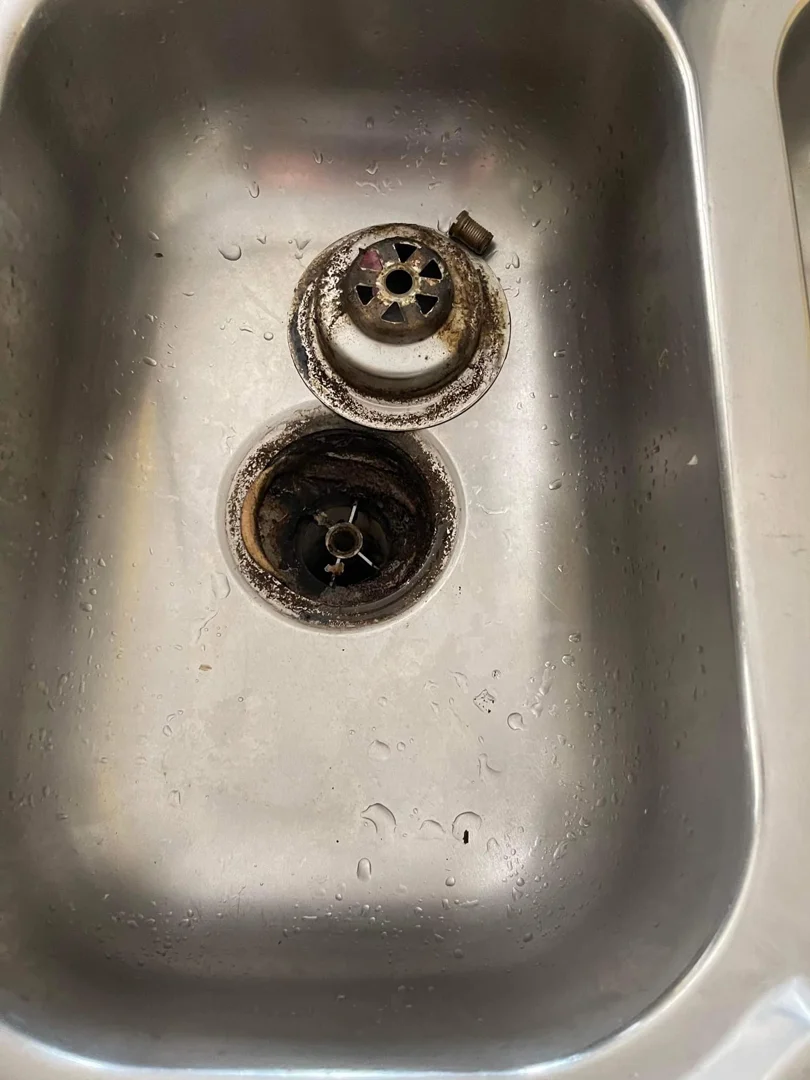 Kitchen sink drain and strainer basket coated in thick black sludge before cleaning.