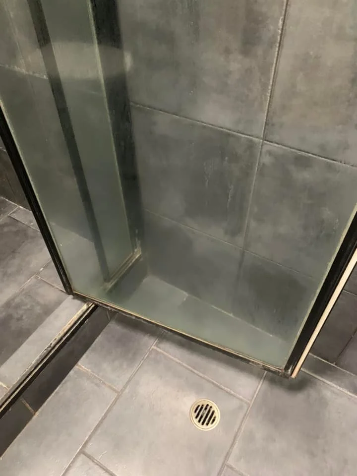 Glass shower door clouded with thick soap scum and hard water mineral deposits before cleaning.