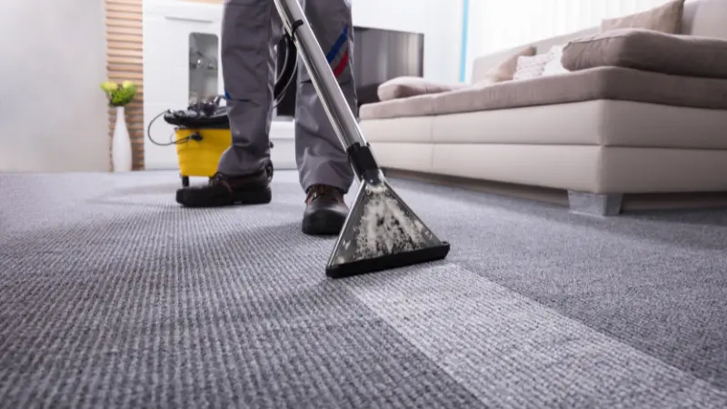 Close-up of a BJR carpet cleaning extraction wand on grey carpet, leaving a visible clean strip behind it next to a yellow industrial machine.