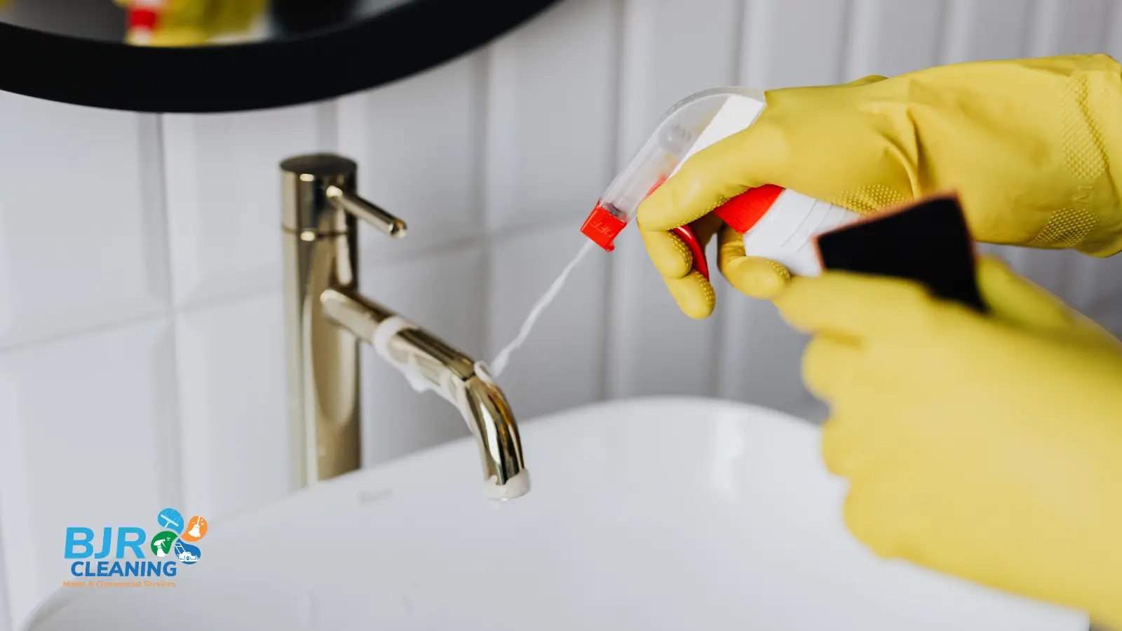 A BJR cleaner wearing yellow rubber gloves spraying foaming cleaner onto a gold faucet in a bathroom with white subway tiles.