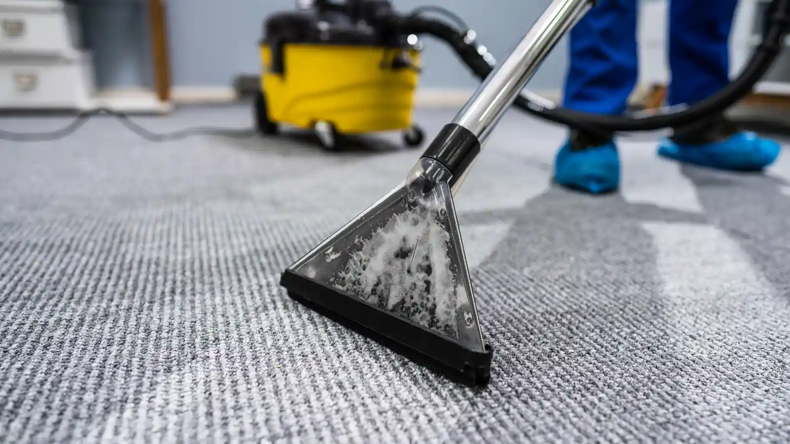 Detailed close-up of a BJR carpet extraction wand with swirling soapy water, alongside a cleaner in blue trousers and a yellow industrial vacuum.