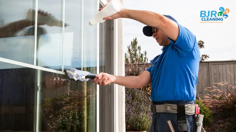 A male BJR window cleaner in a blue t-shirt and cap pouring cleaning solution onto an exterior home window while holding a blue microfiber scrubber.